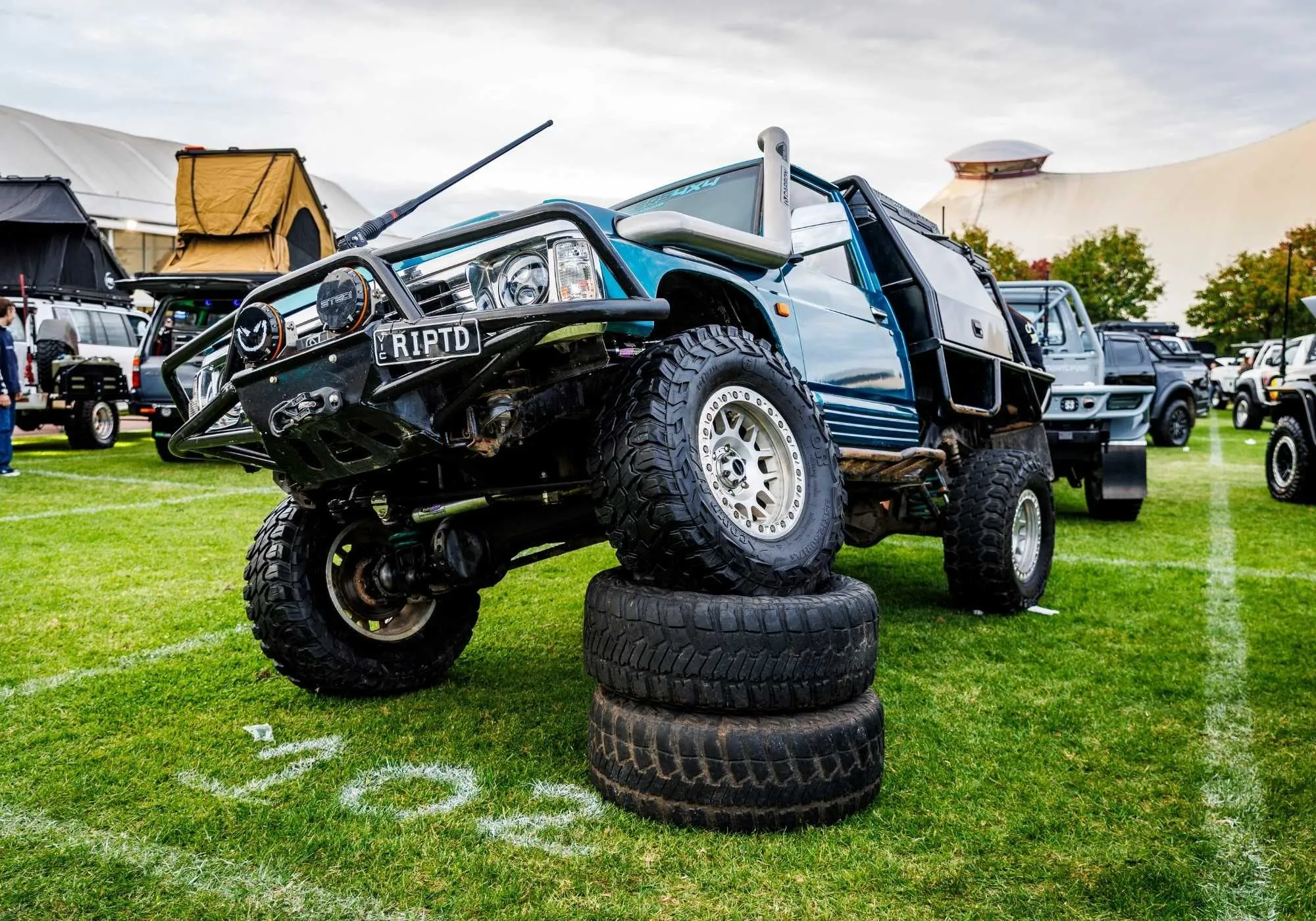 A lifted LandCruiser ute perched on stacked tyres on the grass at Car Builders 4x4 World